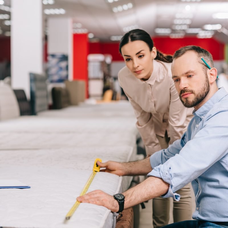 couple measuring mattress with measure tape in furniture store with arranged mattresses