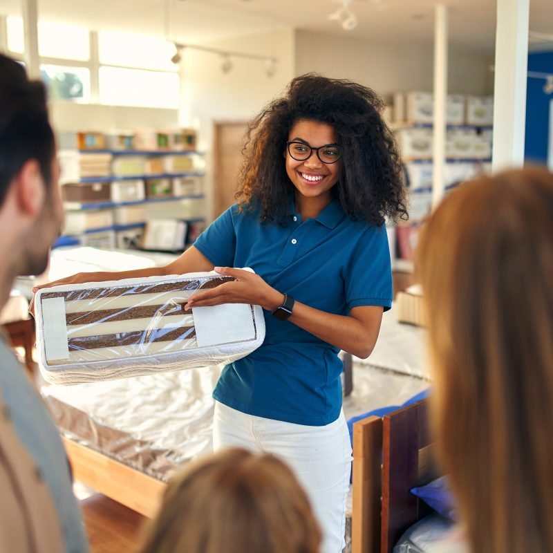An African American woman salesperson consultant consults a young family by showing samples of mattresses in a bed, mattress and pillow store.